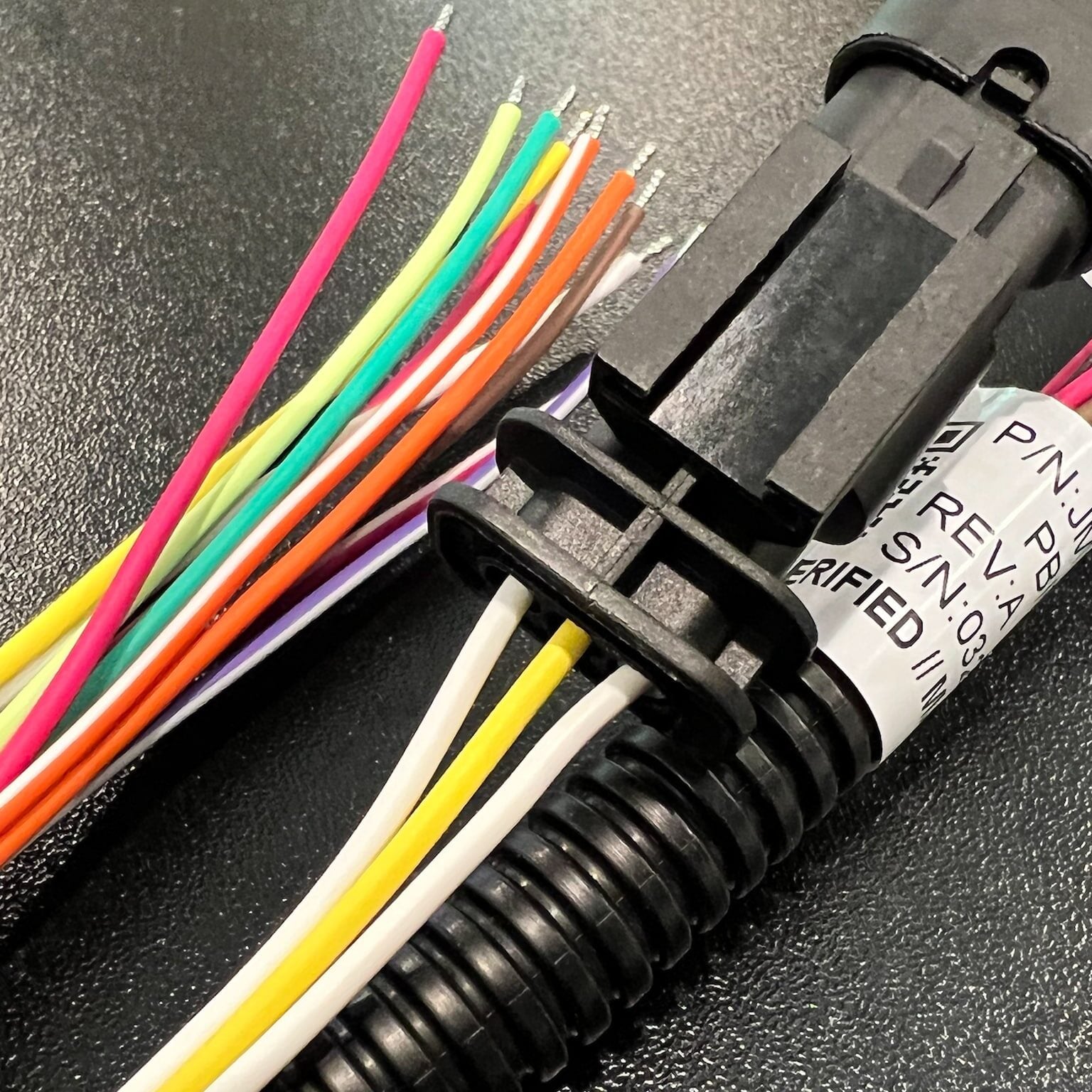 Close-up of a bundle of colorful exposed wires connected to a black plastic connector, with a label attached, resting on a textured black surface.