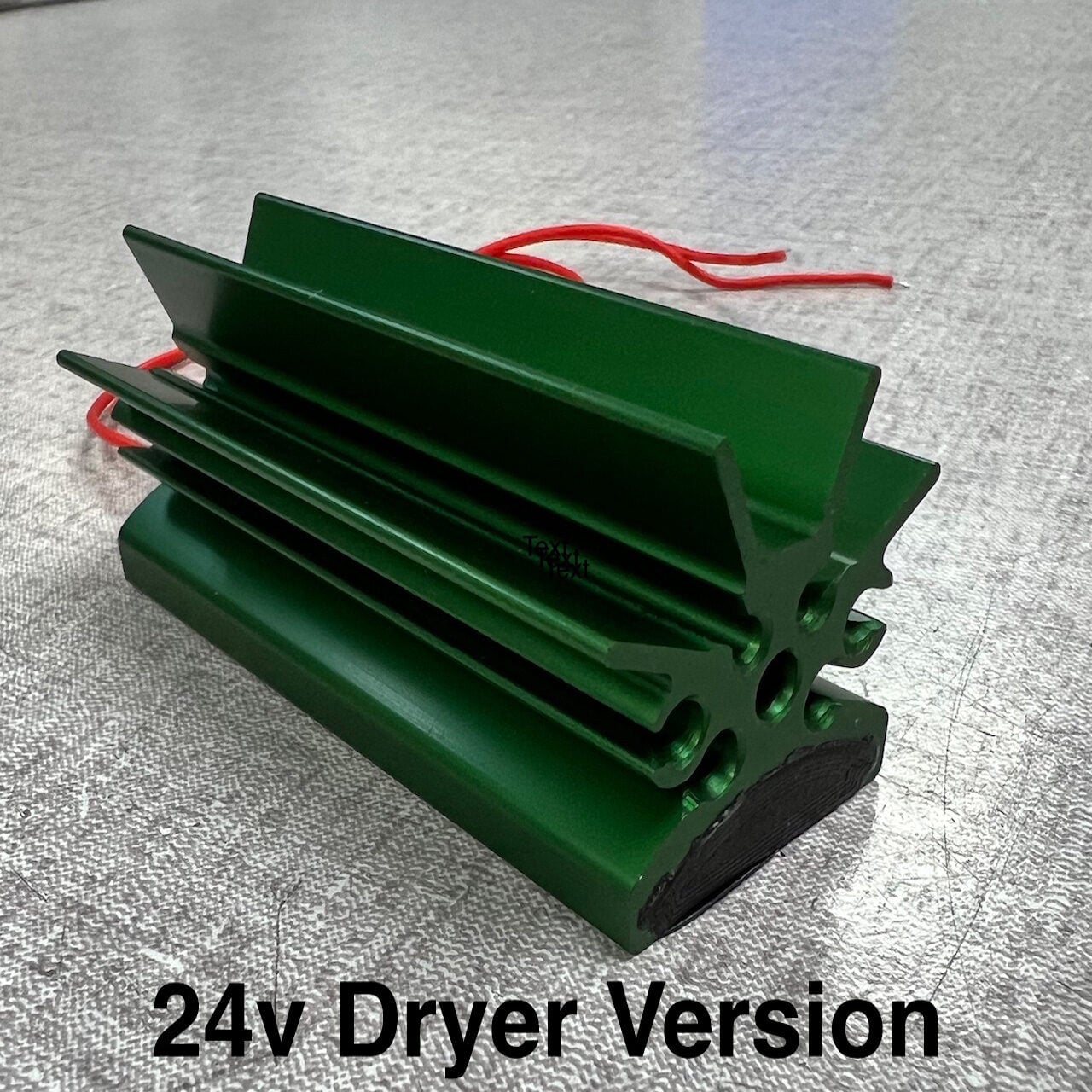A green metallic component with fins and red wires extending from one end is labeled 24v Dryer Version on a gray textured surface.