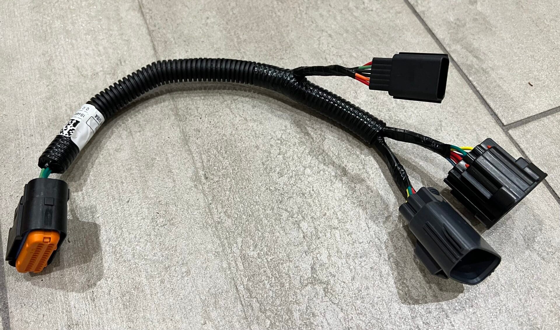 A wiring harness with black plastic connectors and color-coded wires, covered in a black corrugated sleeve, lies on a light-colored tiled floor.