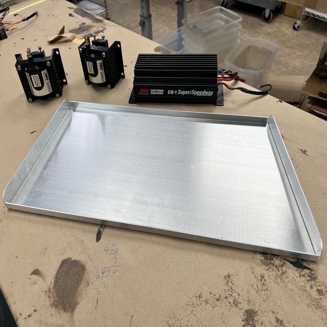 A metal tray sits on a workbench next to two black ignition coils and a CD-1 Super/Speedway ignition control module. Plastic bins and workshop equipment are visible in the background.