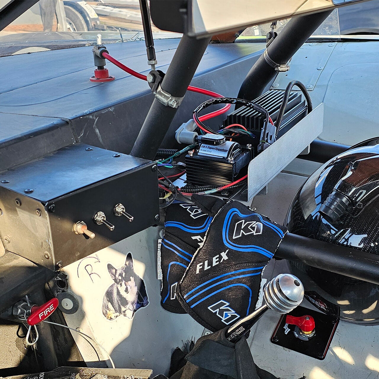 Close-up of a race car’s interior, showing a steering wheel, gear shifter, K1 racing gloves, electronic components, switches, fire extinguisher pull, a helmet, and a sticker of a small dog on the dashboard.